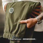 手部展示 Valour Fits 軍綠色梭織機能長褲的布料質感，細節包含圓形貼標與高彈力抽繩腰帶。