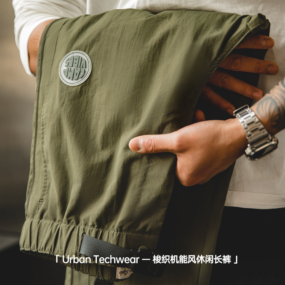 手部展示 Valour Fits 軍綠色梭織機能長褲的布料質感，細節包含圓形貼標與高彈力抽繩腰帶。