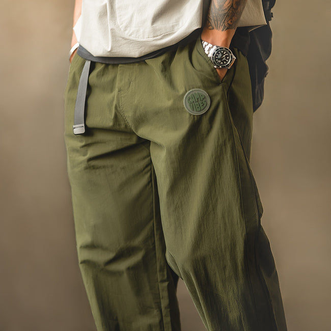 男模特穿著 Valour Fits 軍綠色山系機能工裝褲的近景細節，展示內置快拆織帶腰帶與口袋旁的圓形膠印徽章。 Hover Image